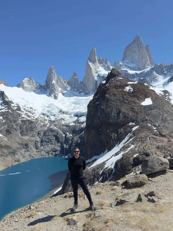 Benedict Grima standing on a rocky ridge above a glacial lake, with snow-covered granite peaks behind